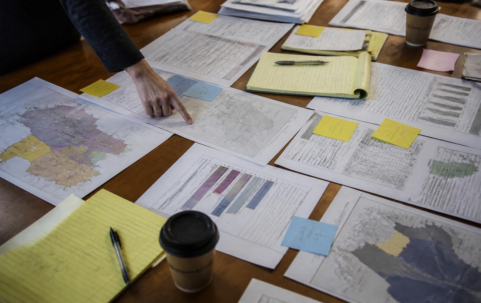 A table covered with state reports, district scorecards, maps, legal pads and highlighted printouts, one hand tracing between two documents.